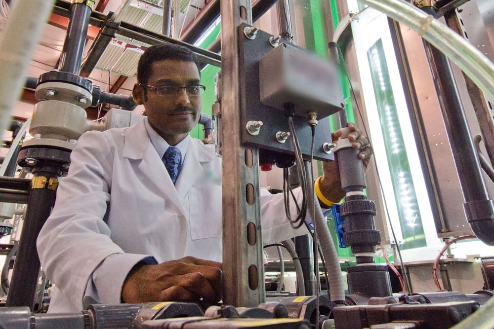 An engineer in glasses and a white lab coat adjusts a valve on a tall system of pipes, tubes and tanks inside a processing plant.
