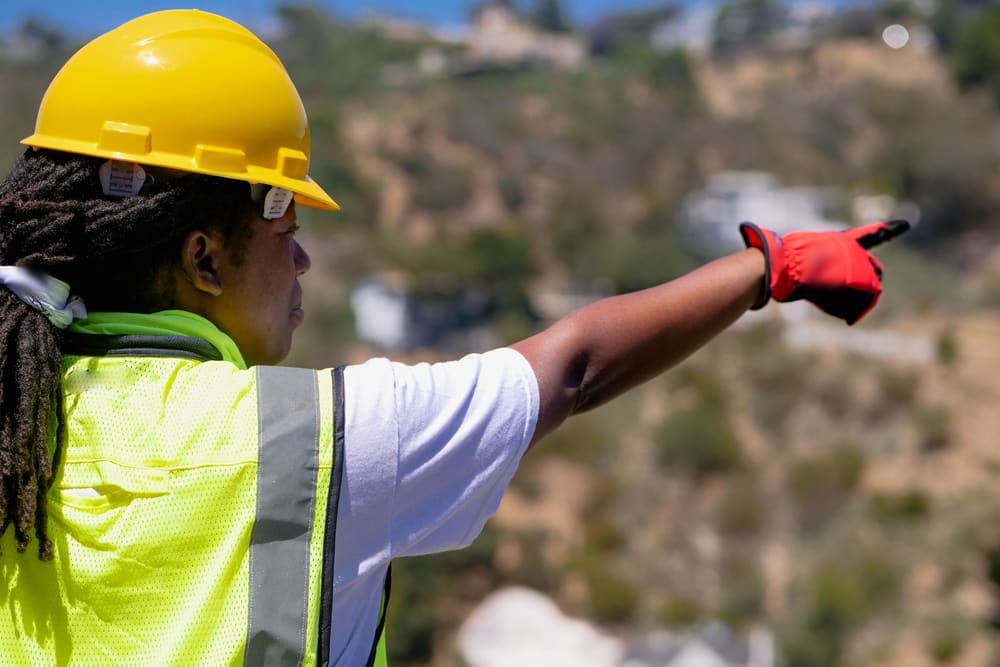 An engineer in a yellow hard hat, a bright safety vest and red work gloves points across a hillside building site.
