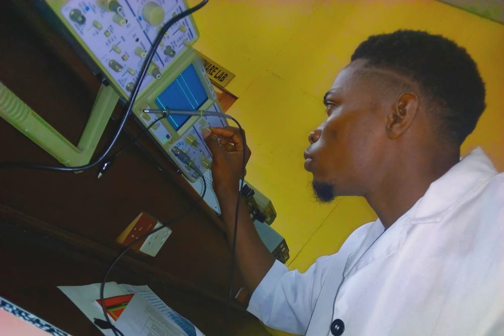 An engineering student in a white lab coat holds a probe to an oscilloscope in a hardware lab, watching the signal on its screen.