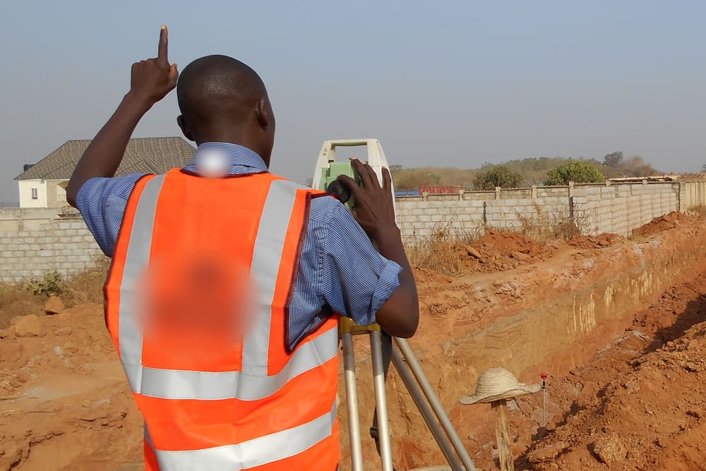 A surveyor in an orange safety vest looks through a measuring instrument on a tripod beside a long trench on a dry, red-earth building site, one hand raised to signal to his team.