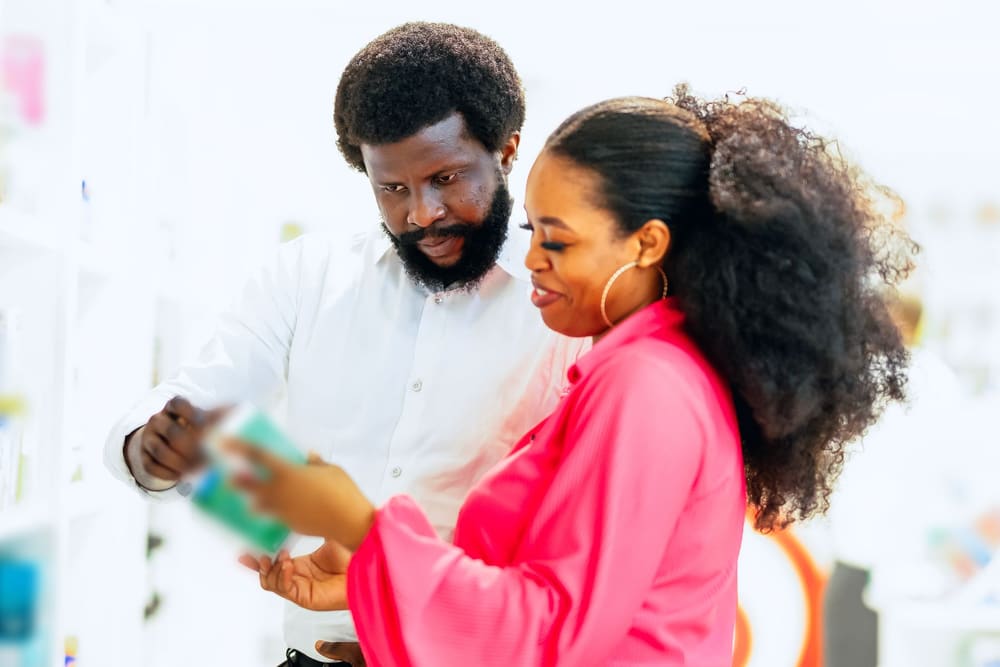In a pharmacy, a man in a white shirt and a woman in a pink blouse look together at a box of medicine taken from the shelves.