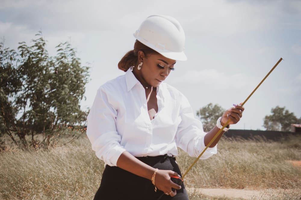 A woman in a white hard hat and white shirt pulls out a long measuring tape on an open, grassy building plot.