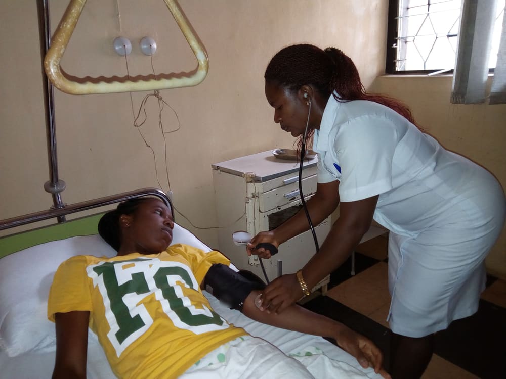 A nurse in a white uniform takes the blood pressure of a young woman lying in a hospital bed, with a cuff on her arm and a stethoscope in her ears.