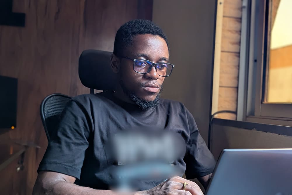 A young man in glasses and a black T-shirt sits in an office chair, concentrating on the laptop in front of him.