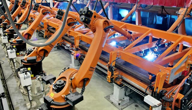 A line of orange robot arms welding the steel side wall of a railway wagon, with sparks at the joints.