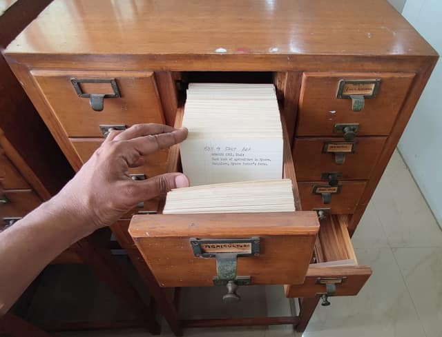 A hand flicks through a drawer of library index cards; each card holds the details of one book.