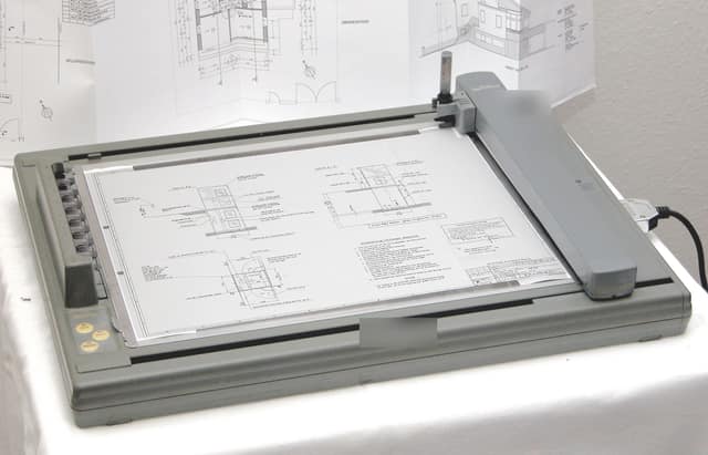 A flatbed pen plotter drawing a large building plan, with more architectural drawings pinned behind it.