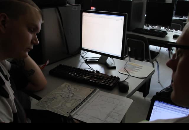 Two people planning a website together at a computer, with a hand-drawn page sketch on the desk.