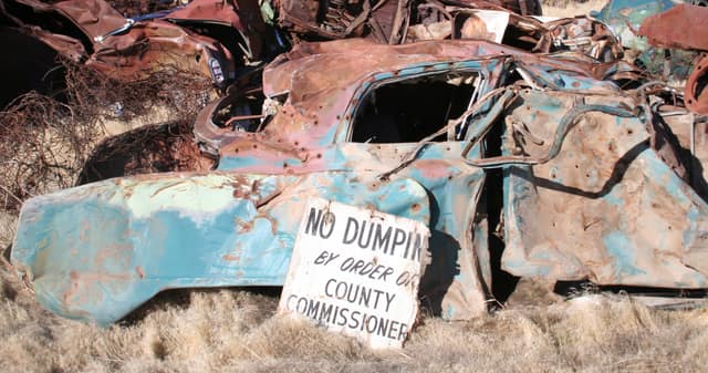 A rusted, dumped car lies in dry grass beside a sign that reads “No dumping by order of County Commissioners”.