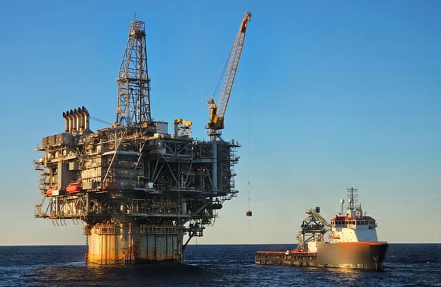 A huge offshore oil and gas platform with a drilling tower and crane stands in the open sea, with a supply ship alongside.