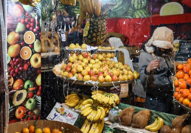 A shopper in a warm hood chooses fruit at a stall piled with apples, oranges, bananas and coconuts under hand-written price signs.