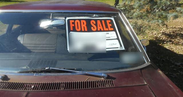 An old red car with a FOR SALE sign stuck inside the windscreen, the details filled in by hand.