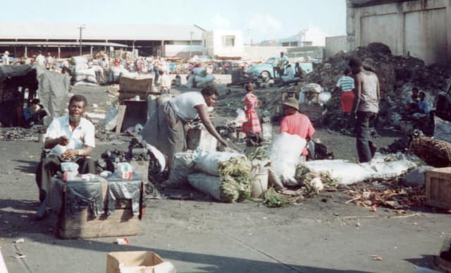 A busy open-air market in downtown Kingston, Jamaica: vendors seated beside sacks and tables of goods, with shoppers moving between them.