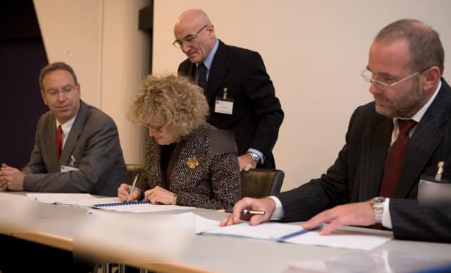 Four business people at a table; two sign copies of a contract while the others watch.