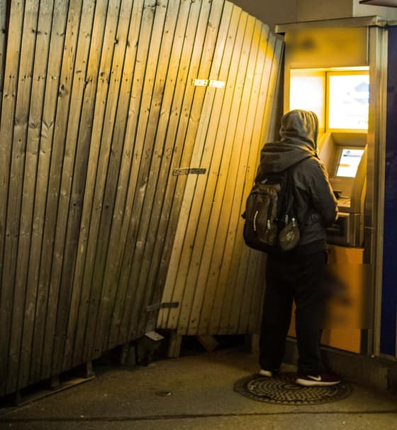A person in a hooded jacket using a cash machine set into a lit wall at night.