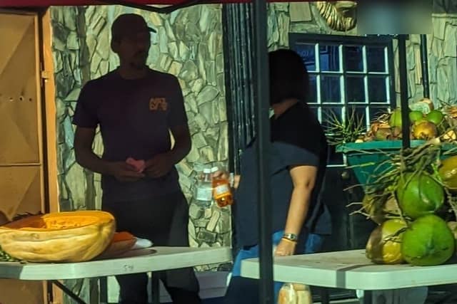 A roadside stall in Trinidad under a red tent: a table piled with green coconuts, another with a pumpkin and a scale, and the vendor standing behind them.