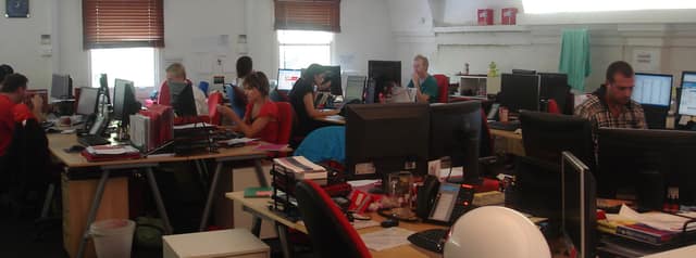 A busy open-plan office: rows of desks with computers, files and phones, and staff at work in each section.