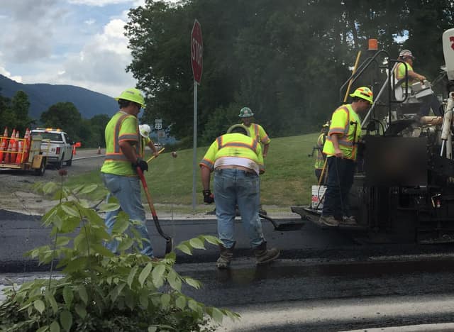 A road crew in high-visibility vests lays fresh asphalt behind a paving machine on a country road.