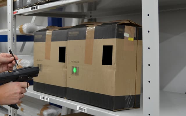 A warehouse worker points a handheld barcode scanner at a box on a shelf to record it in the stock system.