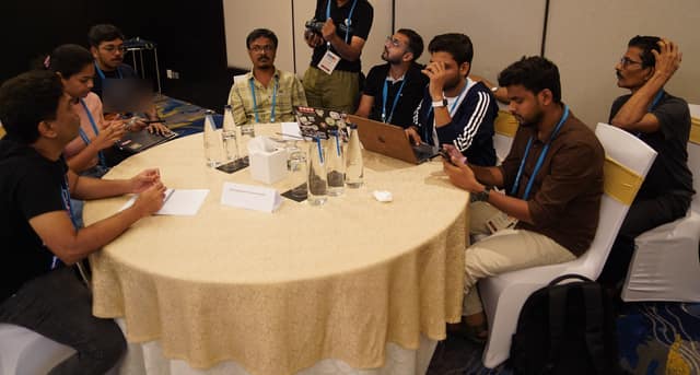 Seven people around a round table with laptops; one stands and listens while the others talk through a problem together.
