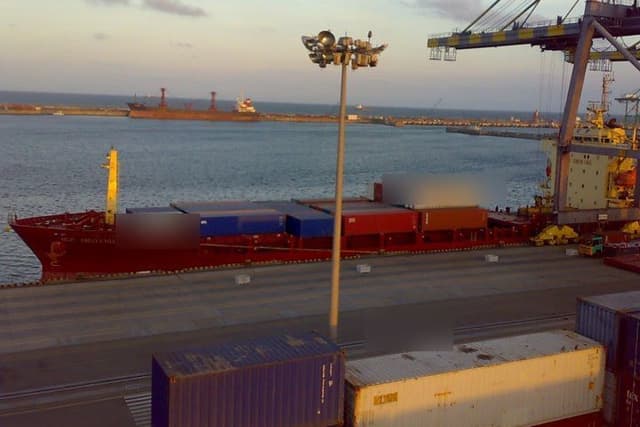 A container ship loaded with containers at a port berth, with a large crane above it and more containers on the quay.