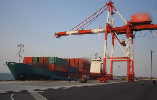 A container ship tied up at a port beside a tall red and white crane that lifts containers on and off.