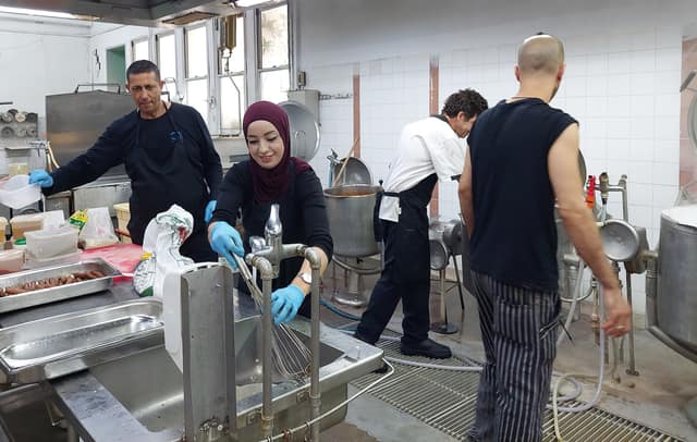 Four cooks at work in a large steel kitchen: one hands over a tub, one whisks at a sink, and two stir big pots.
