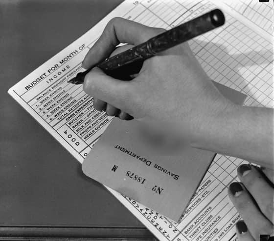 A hand with a fountain pen fills in a printed page headed Budget for month of, with rows for income and food spending, beside a bank savings book.