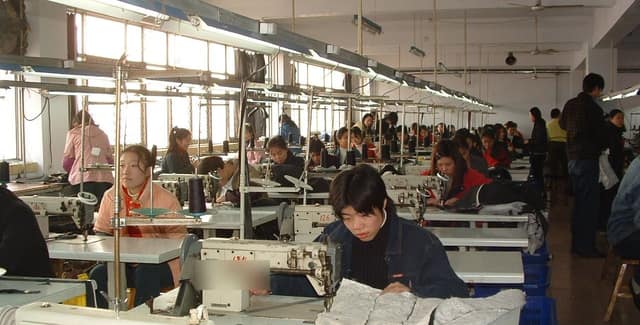 Rows of workers at sewing machines in a garment factory, each stitching pieces of clothing.