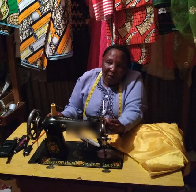 A seamstress sits at a treadle sewing machine in her small workshop, with bright printed cloth hanging behind her.