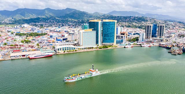 Port of Spain from the air: tall office towers and hotels along the waterfront, the city spreading up green hills, and a work boat crossing the harbour.