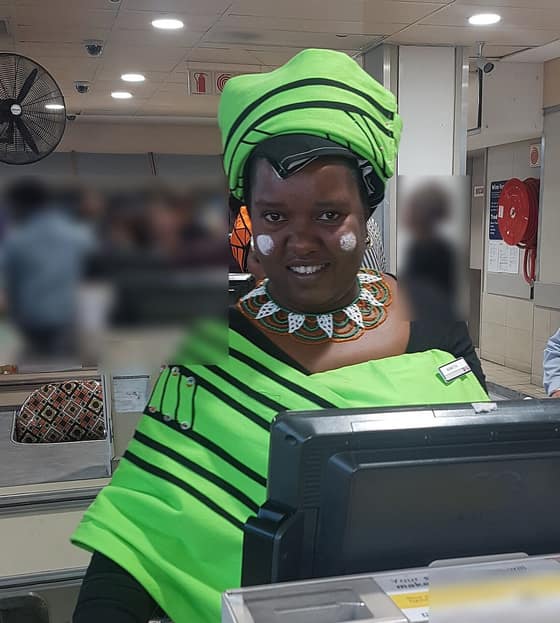 A smiling cashier in a bright green dress and headwrap at a supermarket till, behind the till screen.