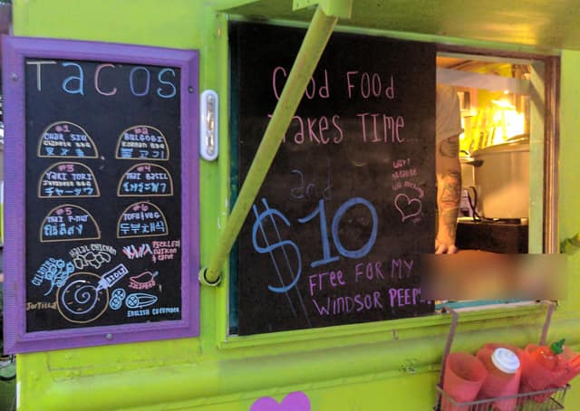 A bright green food truck with a hand-drawn chalkboard menu of tacos and a sign reading Good food takes time, $10.