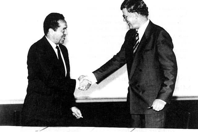 Two men in suits shake hands across a table after reaching an agreement.